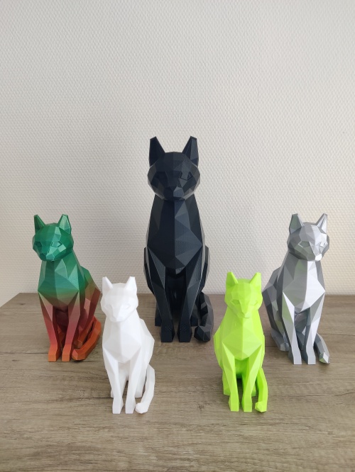 Figurines de chats, pour la SPA de la Mayenne, posées sur un meuble. La plus grande au centre est noire, ensuite une multi couleurs à sa droite puis une blanche un peu plus devant entre les deux. A gauche de la grande figurine noire se trouve un chat gris et devant entre les deux un chat imprimé en 3D vert.