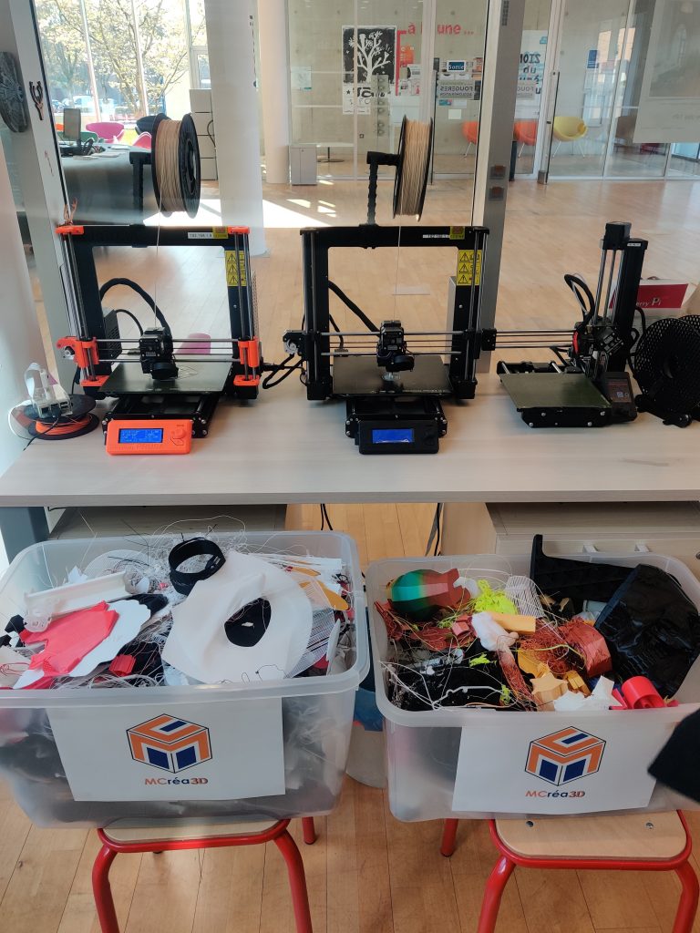 Nous voyons 3 imprimantes filaments posées sur un bureau avec au premier plan, 2 caisses plastiques pleines de filaments PLA, avec le logo MCréa3D dessus.