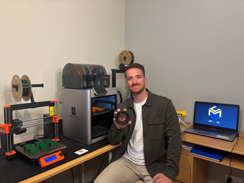 Homme assis tenant une pièce imprimée en 3D. Derrière lui, on retrouve un bureau avec un scanner 3D et son PC ainsi que plusieurs imprimantes filaments